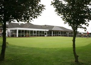 A modern golf clubhouse with large windows overlooks a well-manicured putting green, framed by two trees in the foreground—an ideal setting for a Golf Club AV Project.