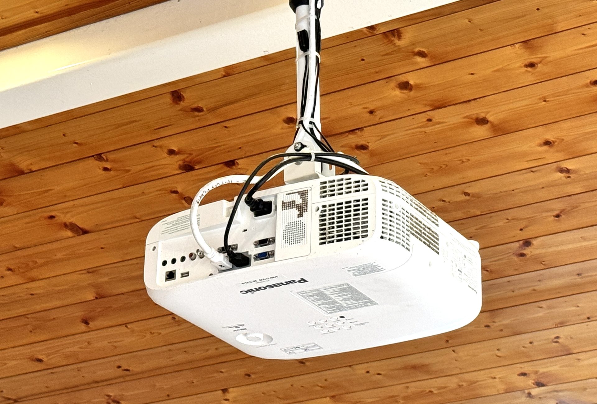 A white Panasonic projector is mounted upside down on a wooden ceiling, connected by several black and white cables as part of a sophisticated Golf Club AV Project.