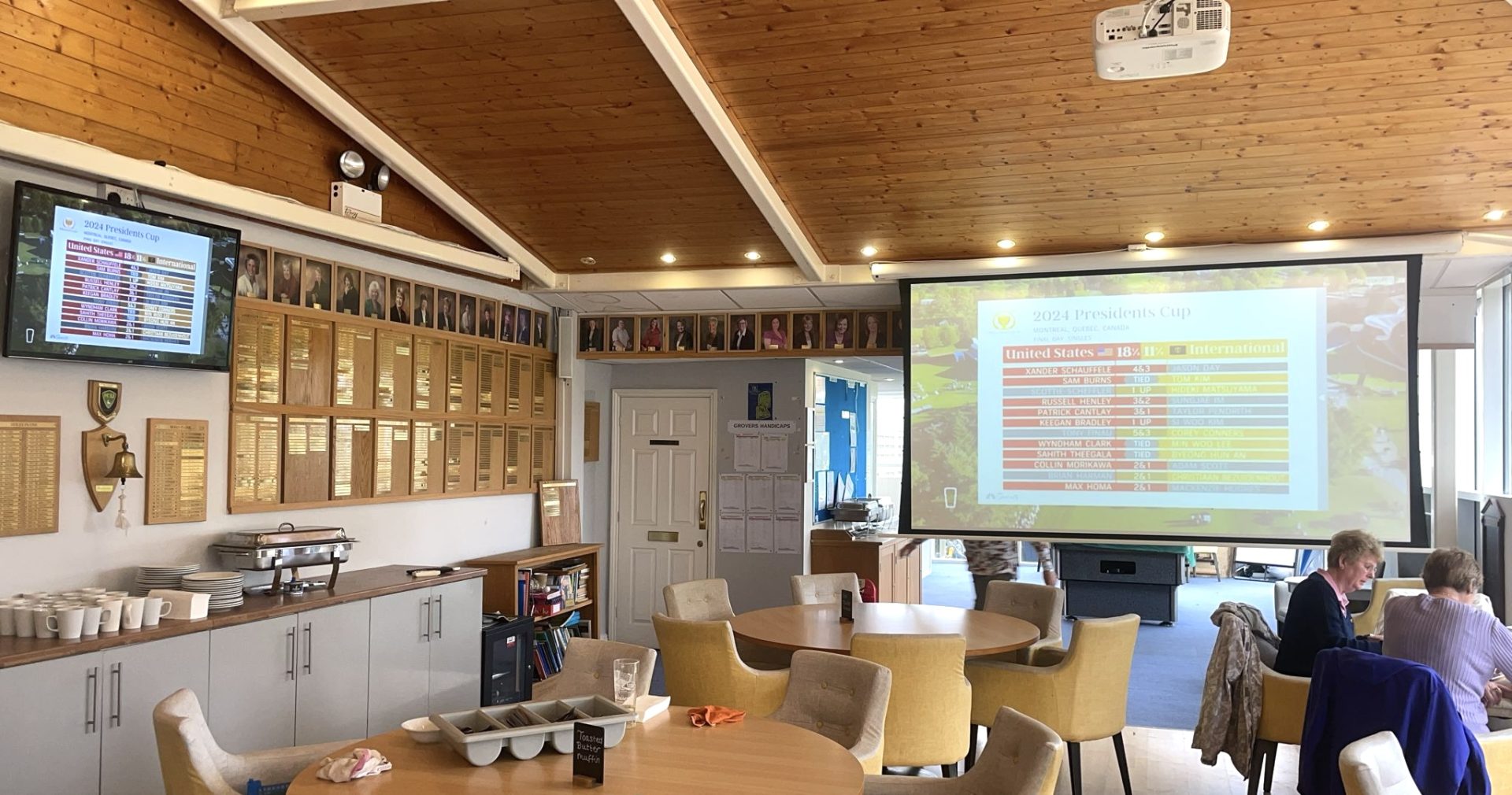 A spacious room with round tables, a buffet set-up on the left, a TV and a projector screen displaying a sports schedule—part of the Golf Club AV Project. Three people sit at a table in the foreground. The wood-panelled ceiling and plaques decorating the walls add to the inviting atmosphere.