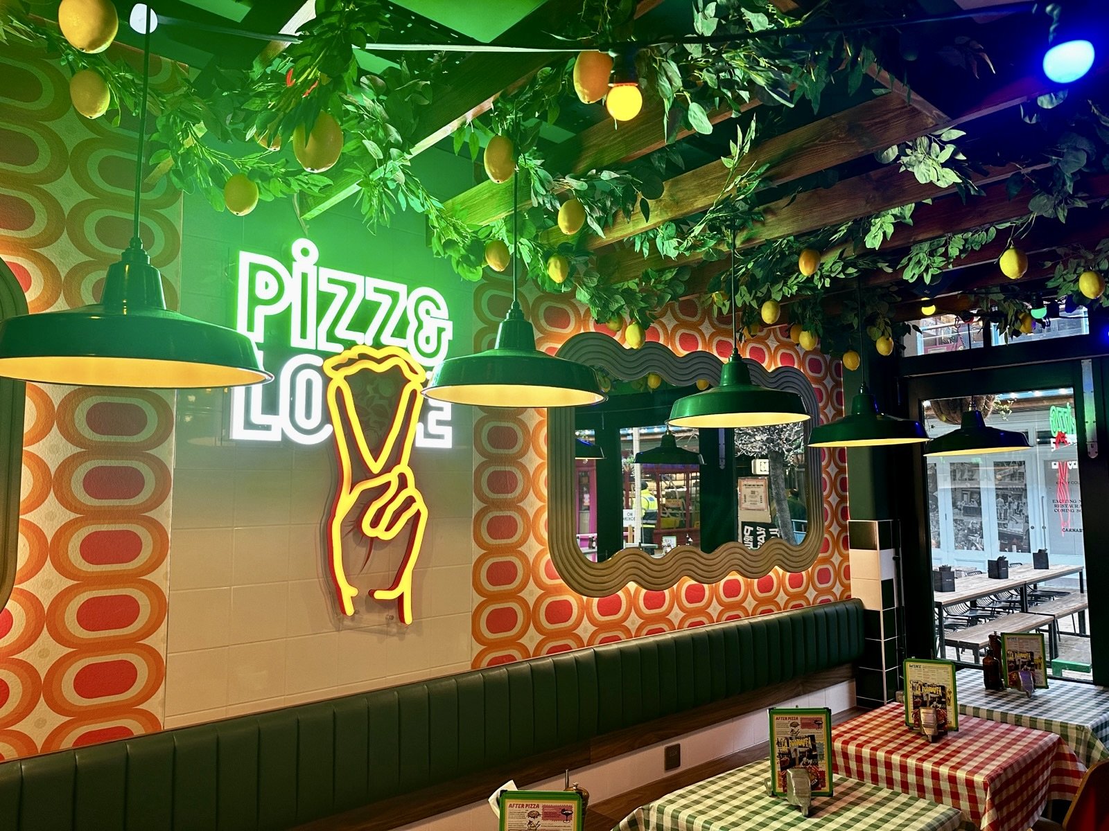 Retro-styled pizza restaurant with a Pizza Love neon sign, hanging lights, leafy ceiling decor, lemons, chequered tablecloths, and outdoor seating visible through the window—a lively space enhanced by vintage audio systems that add to the nostalgic ambience. Retro-styled pizza restaurant with a Pizza Love neon sign, hanging lights, leafy ceiling decor, lemons, chequered tablecloths, and outdoor seating visible through the window—a lively space enhanced by vintage audio systems that add to the nostalgic ambience.