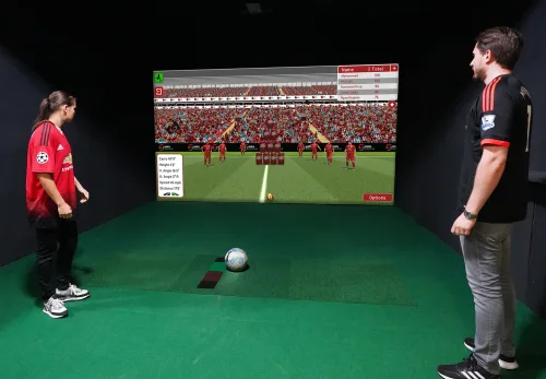 Two people stand on green artificial grass, facing a large video game screen that displays a stadium with virtual football players. With the Lite Edition Sport Simulator, a football sits on the ground between them, ready to be kicked at the screen for an immersive and interactive experience.