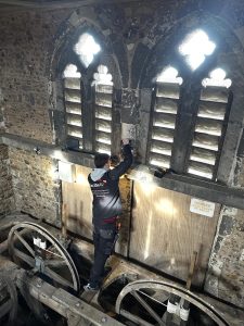 A person in workwear stands near arched windows inside the stone bell tower of Thaxted Church, working on or inspecting equipment as part of a microphone system installation. Large wooden wheels and a Danger Keep Clear sign are visible nearby.