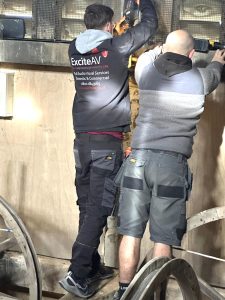Two men stand on a wooden surface working on a wall, possibly as part of a microphone system installation at Thaxted Church. One wears a jacket with Excite AV on the back, while the other wears a grey jacket and shorts. Machinery parts are visible in the foreground.