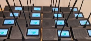 Multiple black tabletop microphone units with screens displaying the same logo are arranged in rows, ready for use in the Uttlesford Council Chambers. The devices have long aerials and are positioned closely together on a light-coloured surface.