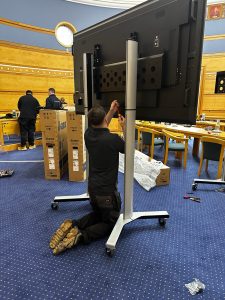 A person kneels on the floor assembling a large TV on a stand in a conference room in the Uttlesford Council Chambers, with tools and packaging about. Two others work in the background.