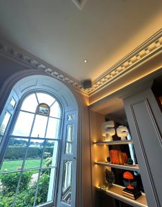 A tall arched window with a stained glass detail overlooks a green landscape, evoking the serene charm of St Albans. Inside, built-in shelves hold books, abstract sculptures, and an orange lamp under elegant ceiling mouldings with warm lighting—reminiscent of a stylish cocktail bar at Sopwell House.