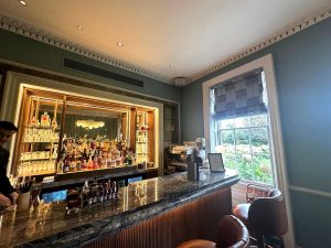 A stylish cocktail bar at Sopwell House in St Albans, featuring a marble counter, shelves filled with bottles, a barista machine, and a window letting in natural light. Brown bar stools and greenery are visible outside the window.