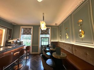 A stylish café in St Albans with green panelled walls, brown leather seating, round tables, modern light fittings, bar stools, and large windows letting in natural light—perfect for relaxing or enjoying a signature creation from the cocktail bar.