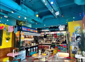 A vibrant diner in Cardiff with colourful decor, hanging lights, mismatched chairs, wall art, and a counter serving delicious pizza and drinks. Various signs and shirts hang above the bar area at Pizza Pilgrims, creating a lively, playful atmosphere.