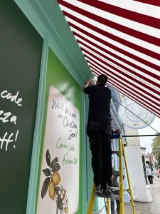 A person stands on a ladder under a red and white striped canopy, working on a shopfront with green panels—one of which reads, Coming Late Autumn 24 and We’ll be here in Chelsea soon! The scene hints at the arrival of Pizza Pilgrims bringing their much-loved pizza to Chelsea. A person stands on a ladder under a red and white striped canopy, working on a shopfront with green panels—one of which reads, Coming Late Autumn 24 and We’ll be here in Chelsea soon! The scene hints at the arrival of Pizza Pilgrims bringing their much-loved pizza to Chelsea.
