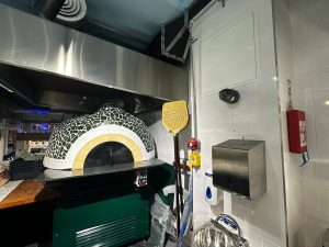 A tiled pizza oven with a yellow paddle stands in a commercial kitchen at this Chelsea restaurant. Nearby are a metal paper towel dispenser, a handwashing basin, and a red fire extinguisher mounted on the white tiled wall—reflecting the signature set-up found at Pizza Pilgrims’ popular Chelsea pizza spot. A tiled pizza oven with a yellow paddle stands in a commercial kitchen at this Chelsea restaurant. Nearby are a metal paper towel dispenser, a handwashing basin, and a red fire extinguisher mounted on the white tiled wall—reflecting the signature set-up found at Pizza Pilgrims’ popular Chelsea pizza spot.
