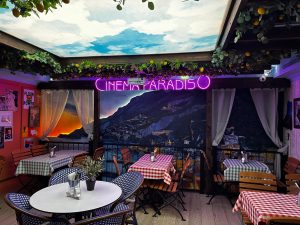 A cosy café with chequered tablecloths, potted plants, wall art, and a mural of a coastal town at sunset. CINEMA PARADISO glows in pink neon above a faux balcony with greenery and a sky ceiling—making this Chelsea restaurant the perfect spot for Pizza Pilgrims to gather and enjoy an inviting atmosphere. A cosy café with chequered tablecloths, potted plants, wall art, and a mural of a coastal town at sunset. CINEMA PARADISO glows in pink neon above a faux balcony with greenery and a sky ceiling—making this Chelsea restaurant the perfect spot for Pizza Pilgrims to gather and enjoy an inviting atmosphere.