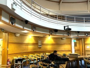 A person sits at a table with equipment in a wood-panelled room with yellow chairs, curved balcony, wall speakers, and exit signs visible; the setting closely resembles the Council Chambers in Uttlesford.