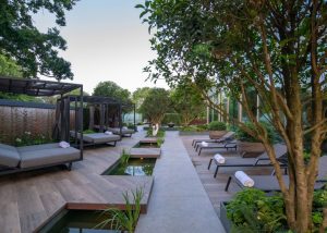 Modern outdoor spa area with loungers, cabanas, and small ponds surrounded by lush green trees and plants. Discreetly integrated Garden Audio Systems provide soothing background music as rolled towels rest on the chairs next to the walkway.