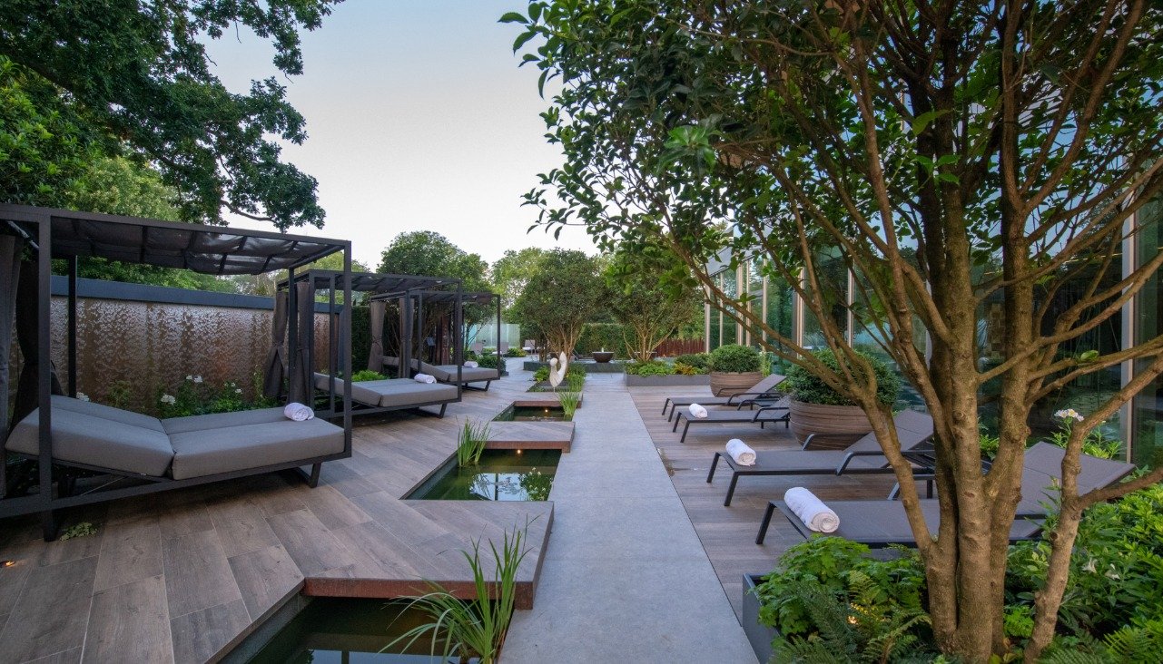 Modern outdoor spa area with loungers, cabanas, and small ponds surrounded by lush green trees and plants. Discreetly integrated Garden Audio Systems provide soothing background music as rolled towels rest on the chairs next to the walkway.