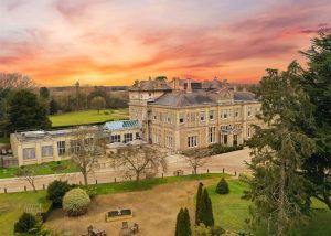 A large historic mansion with ornate architecture, reminiscent of the elegant Down Hall Hotel in Essex, is surrounded by trees and gardens under a vibrant, colourful sunset sky.