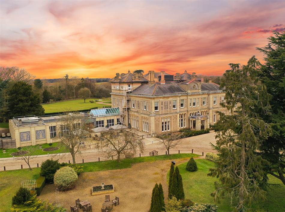 A large historic mansion with ornate architecture, reminiscent of the elegant Down Hall Hotel in Essex, is surrounded by trees and gardens under a vibrant, colourful sunset sky.
