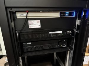 A close-up of audio and network equipment in a black server rack at Builder Depot in New Southgate, featuring an amplifier and control units with indicator lights, labels, and visible cables—essential building supplies for seamless connectivity.