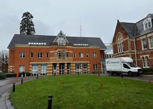 A red-brick building with white-trimmed windows stands beside a smaller, older brick structure that resembles historic Uttlesford Council Chambers. A white van labelled Interiors is parked on a driveway, and small red flags mark spots on the green lawn in front.
