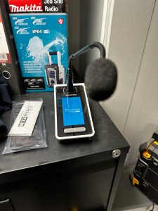 A smartphone with a large external microphone attached is displaying the Atmosphere app, sitting on a black cupboard alongside building supplies and tools, with a Makita site radio box—capturing the busy atmosphere of Builder Depot in New Southgate.