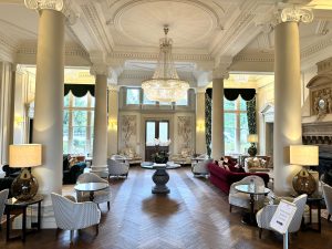 Elegant hotel lounge at Down Hall Hotel in Essex with tall columns, ornate ceiling, large chandelier, classic furniture, and large windows letting in natural light. The room has a warm, inviting atmosphere with decorative lamps and greenery outside.