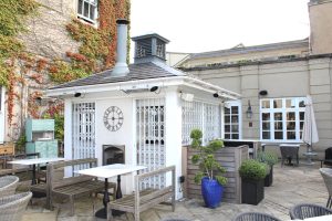 Outdoor patio at the Downhall Hotel in Essex featuring a small white building, benches, tables, and potted plants. The building has a chimney, clock, and elegant white lattice gates. Ivy covers one wall; wicker chairs and a blue pot decorate the inviting area.