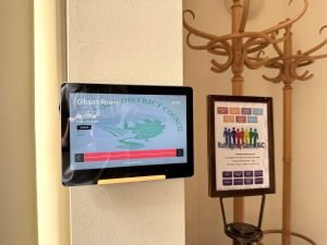 A wall-mounted display shows Gibson Room and council graphics, reflecting the official style of Uttlesford Council. Next to it is a framed sign for the Multi-Agency Centre on a wooden coat rack with multiple hooks, characteristic of the Council Chambers in Uttlesford.