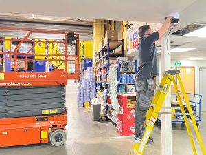 A man stands on a yellow ladder, using a power tool on a ceiling beam in a warehouse at Builder Depot New Southgate. Nearby are shelves with supplies and an orange scissor lift. The area is brightly lit and organised.
