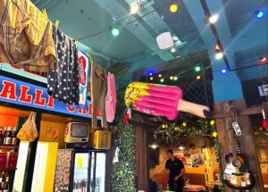 A quirky interior with colourful fairy lights, clothes hanging from the ceiling, a large inflatable ice cream, retro telly, guitar on a wall, and a person standing in a cosy Cardiff restaurant or café—perfect for grabbing a slice at Pizza Pilgrims.