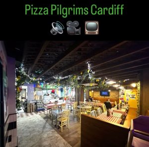 A cosy pizzeria with chequered tablecloths, hanging lights, and greenery, Pizza Pilgrims Cardiff offers the perfect atmosphere to enjoy delicious pizza. Above the inviting dining area, icons of a speaker, camera, and TV add a modern touch to this charming Cardiff hotspot.