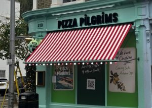 A mint green shopfront with a red and white striped awning reads Pizza Pilgrims. Festive lights twinkle above signs and a QR code, creating a welcoming atmosphere in Chelsea. A decorated tree stands outside on the left, adding charm to this vibrant spot.