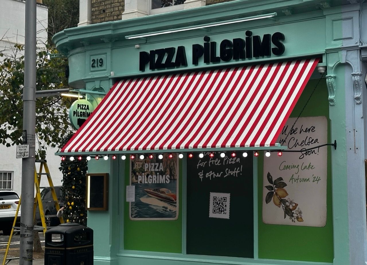 A mint green shopfront with a red and white striped awning reads Pizza Pilgrims. Festive lights twinkle above signs and a QR code, creating a welcoming atmosphere in Chelsea. A decorated tree stands outside on the left, adding charm to this vibrant spot. A mint green shopfront with a red and white striped awning reads Pizza Pilgrims. Festive lights twinkle above signs and a QR code, creating a welcoming atmosphere in Chelsea. A decorated tree stands outside on the left, adding charm to this vibrant spot.