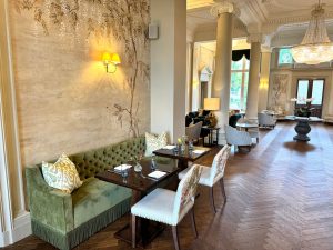 Elegant dining area at the Downhall Hotel in Essex, featuring a green velvet bench, floral-patterned chairs, laid table, ornate wall designs, and large windows letting in natural light. Chandeliers and tall columns add to the luxurious atmosphere.