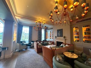 Elegant lounge at Sopwell House in St Albans with large windows, comfy sofas, armchairs, chandeliers, bookshelves with warm lighting, and a fireplace. Daylight streams in, creating a cosy and inviting atmosphere—perfect for relaxing or enjoying drinks from the cocktail bar.