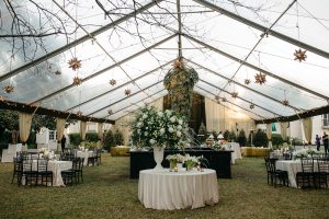 Elegant outdoor wedding reception under a clear marquee with fairy lights, hanging star decorations, round tables with white linen, black chairs, and large floral centrepieces on green grass. Seamlessly integrated Garden Audio Systems ensure that music and speeches are perfectly enjoyed throughout the celebration.