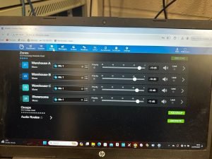 A computer screen displays an audio control interface with four zones: Warehouse A, Warehouse B, Warehouse C, and Showrooms at Builder Depot New Southgate, each with volume and priority sliders.