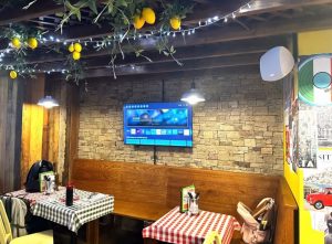A cosy restaurant in Cardiff with wooden decor, chequered tablecloths, a TV on a brick wall, and artificial lemons and vines hanging from the ceiling. Two small tables are set for diners, capturing the inviting atmosphere of Pizza Pilgrims.