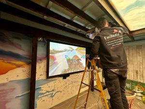 A person on a ladder installs a projector in a room with colourful wall art, reminiscent of the vibrant atmosphere at Pizza Pilgrims Chelsea. The projector displays a motorcycle road view while the installer, wearing a black ExciteAV jacket, works carefully to perfect the setup. A person on a ladder installs a projector in a room with colourful wall art, reminiscent of the vibrant atmosphere at Pizza Pilgrims Chelsea. The projector displays a motorcycle road view while the installer, wearing a black ExciteAV jacket, works carefully to perfect the setup.