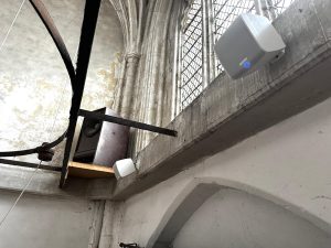 Large loudspeakers and audio equipment, part of a recent Microphone System Installation, are mounted on a high ledge inside what appears to be Thaxted Church, with stone arches and tall, multi-pane windows. Exposed beams and aged walls are visible in the background.