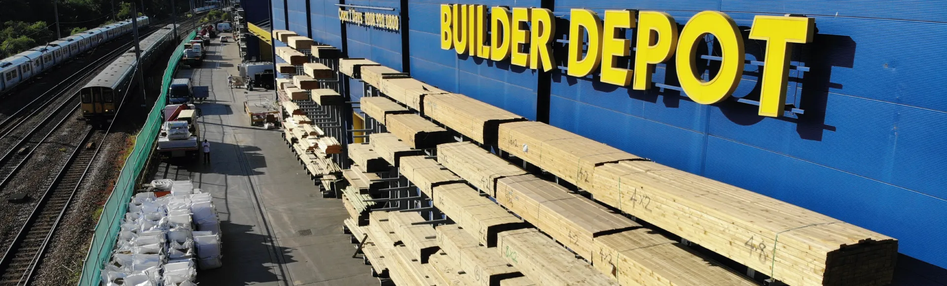A large hardware shop with yellow BUILDER DEPOT signage on a blue wall, located in New Southgate, showing stacks of timber and building supplies next to railway tracks.