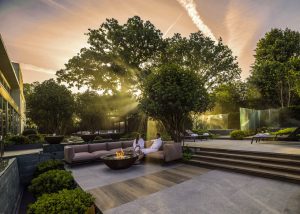 Two people in white robes sit by a fire pit on an outdoor patio surrounded by trees and loungers, with sunlight streaming through the branches at sunset. Discreet Garden Audio Systems provide a soothing soundtrack to this tranquil scene, enhancing the ambience of their peaceful retreat.
