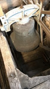 Large, weathered metal bell with inscriptions, mounted in a wooden and metal frame, viewed from above inside the rustic wooden bell tower of Thaxted Church—a historic setting where modern additions such as microphone system installation discreetly complement its timeless atmosphere.