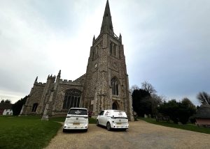 A historic stone church with a tall steeple—Thaxted Church—stands proudly with two white vans parked in front on a gravel path, surrounded by grass and trees under an overcast sky, possibly there for a microphone system installation.