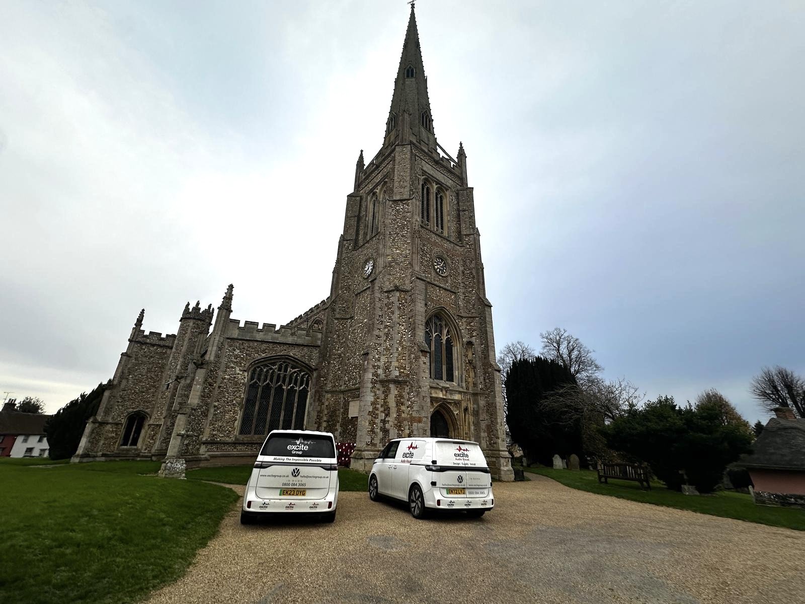 A historic stone church with a tall steeple—Thaxted Church—stands proudly with two white vans parked in front on a gravel path, surrounded by grass and trees under an overcast sky, possibly there for a microphone system installation.