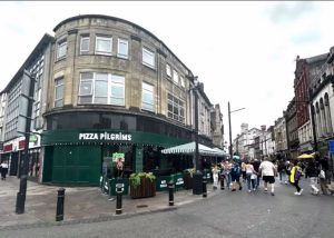 A busy street scene in Cardiff with people walking past a corner building housing Pizza Pilgrims under a cloudy sky. The area features historic buildings, pedestrians, outdoor seating, and the inviting aroma of freshly baked pizza.