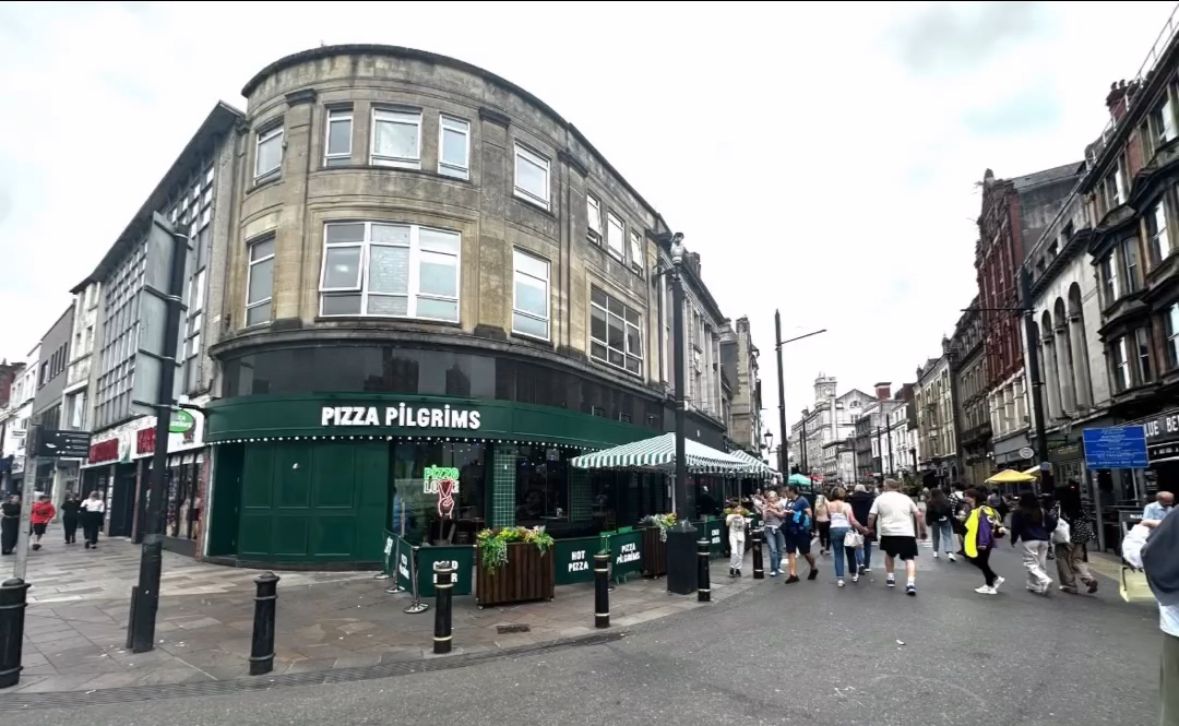 A busy street scene in Cardiff with people walking past a corner building housing Pizza Pilgrims under a cloudy sky. The area features historic buildings, pedestrians, outdoor seating, and the inviting aroma of freshly baked pizza.