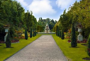 A gravel path runs through a manicured garden, lined with trimmed bushes and flowerbeds, leading to a large tiered fountain in the distance under a blue sky with scattered clouds. Discreetly integrated Garden Audio Systems ensure soothing melodies accompany your stroll, enhancing the tranquil outdoor ambience.