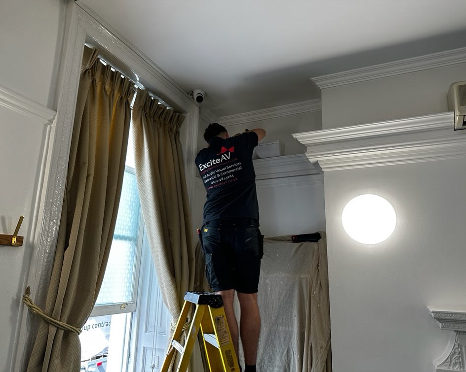 A person stands on a ladder installing or adjusting a device near the ceiling in a room with cream curtains and white walls. The person wears a dark Excite AV shirt and shorts, working as part of an installation project for Mayfair Islamic Centre. A person stands on a ladder installing or adjusting a device near the ceiling in a room with cream curtains and white walls. The person wears a dark Excite AV shirt and shorts, working as part of an installation project for Mayfair Islamic Centre.