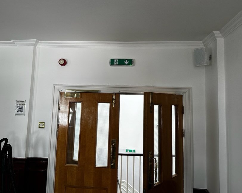 Two open wooden doors with glass panels lead to a lit corridor within the Mayfair Islamic Centre. An emergency exit sign with a downward arrow is above the doors on a white wall, while a fire alarm and a small framed notice are also visible—a scene that could serve as an insightful case study for safety features in contemporary Islamic Centre design. Two open wooden doors with glass panels lead to a lit corridor within the Mayfair Islamic Centre. An emergency exit sign with a downward arrow is above the doors on a white wall, while a fire alarm and a small framed notice are also visible—a scene that could serve as an insightful case study for safety features in contemporary Islamic Centre design.