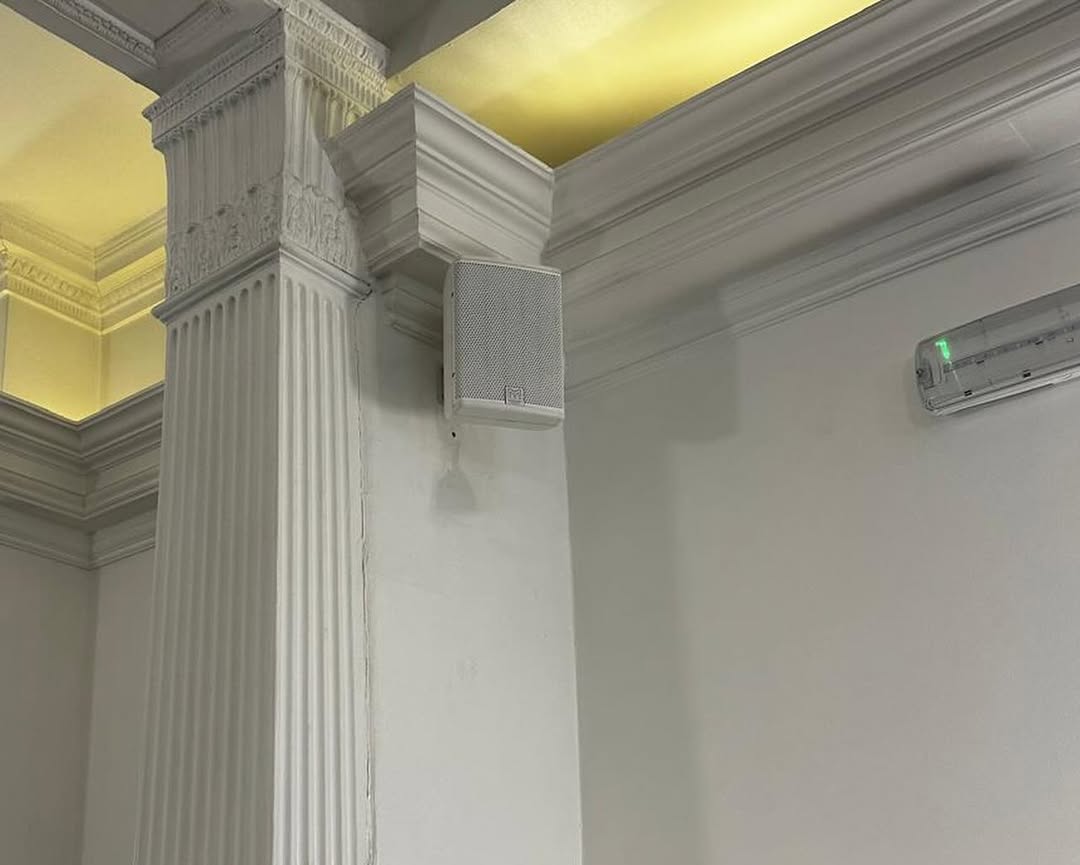A white loudspeaker is mounted high on a white wall near a decorative pillar at the Mayfair Islamic Centre. An air conditioning unit is installed on the right side of the image, while soft yellow lighting highlights the ceiling’s moulding details, creating an inviting atmosphere within this Islamic centre. A white loudspeaker is mounted high on a white wall near a decorative pillar at the Mayfair Islamic Centre. An air conditioning unit is installed on the right side of the image, while soft yellow lighting highlights the ceiling’s moulding details, creating an inviting atmosphere within this Islamic centre.